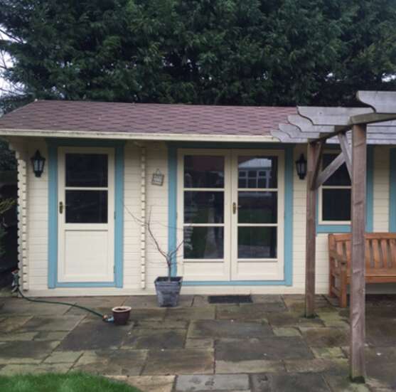 Tiger Delta Log Cabin painted in cream and blue on garden patio with wooden bench with shingle roof