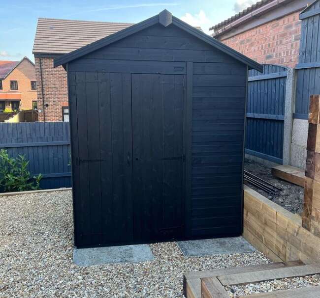 A picture containing a Tiger Overlap Apex Garden Shed painted in black on patio with painted garden fence and shrubs