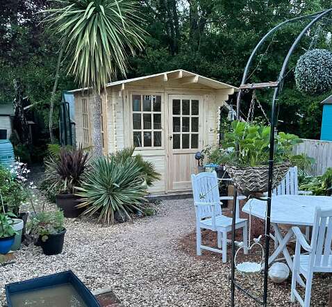 Tiger Sheds Caspian Log Cabin in pebbled garden with palm tree, potted plants, garden arch and table and chairs