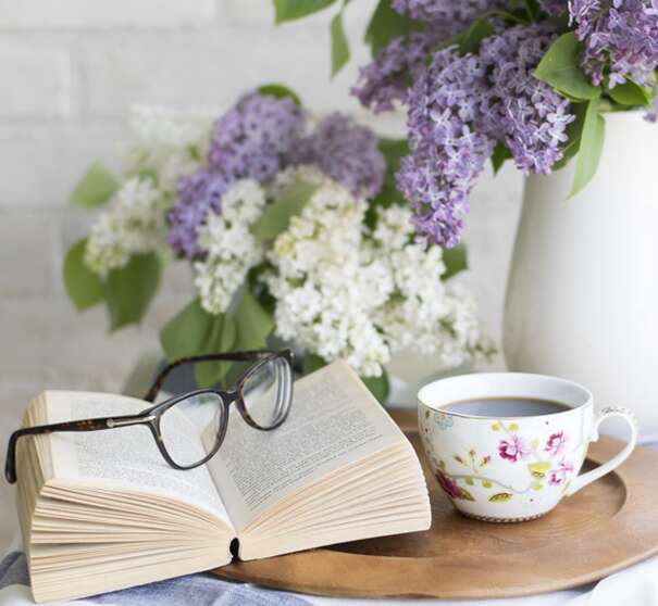 A picture of an opening book with glasses placed on top, a floral china cup of coffee and purple and white flowers in  vase on wooden tray