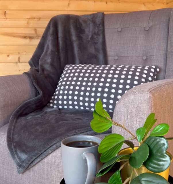 A picture of a cosy reading corner in a log cabin with grey armchair, fur throw, mug of coffee and plant
