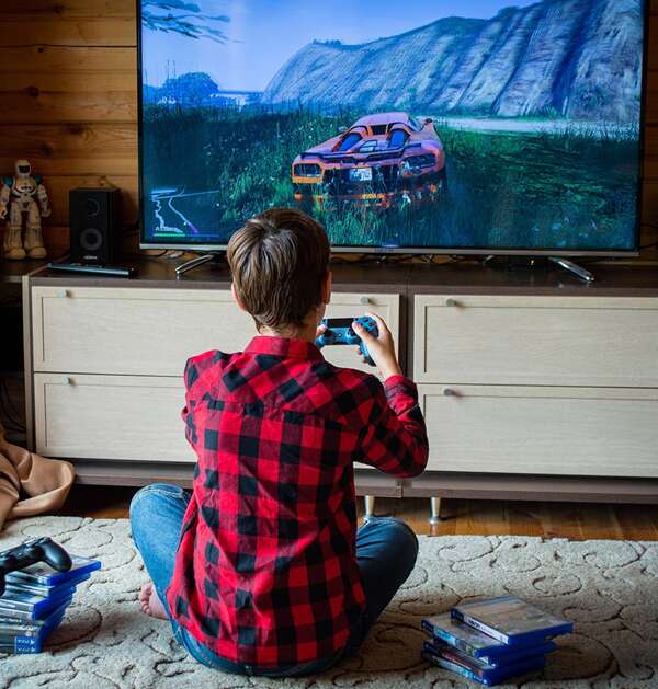 A picture containing a boy playing video games sat on floor rug looking up a the tv, garden games room
