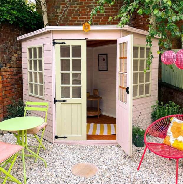 A picture containing the Tiger Corner Summerhouse painted in soft pink and cream in gravelled garden with bright pink and lime green metal garden furniture