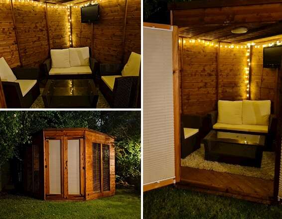 A picture containing the Tiger Vista Corner Summerhouse with white slatted window blinds, comfy garden furniture inside with cream rug and coffee table in garden at night