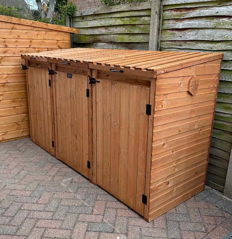 A picture containing a triple wooden bin store on paved patio next to garden fence