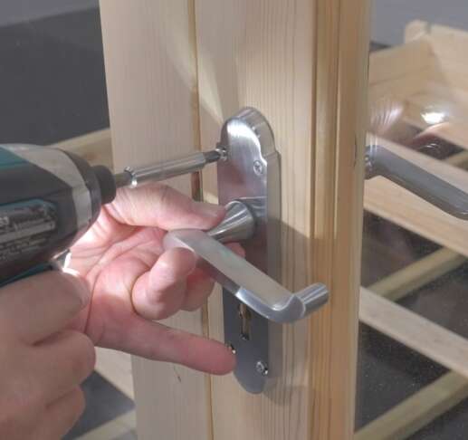 A hand holding a screwdriver and attaching a stainless steel door handle to a log cabin door